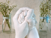 White plaster sculpture of two hands holding each other, flanked by glass vases with small white flowers tied with light blue ribbons.