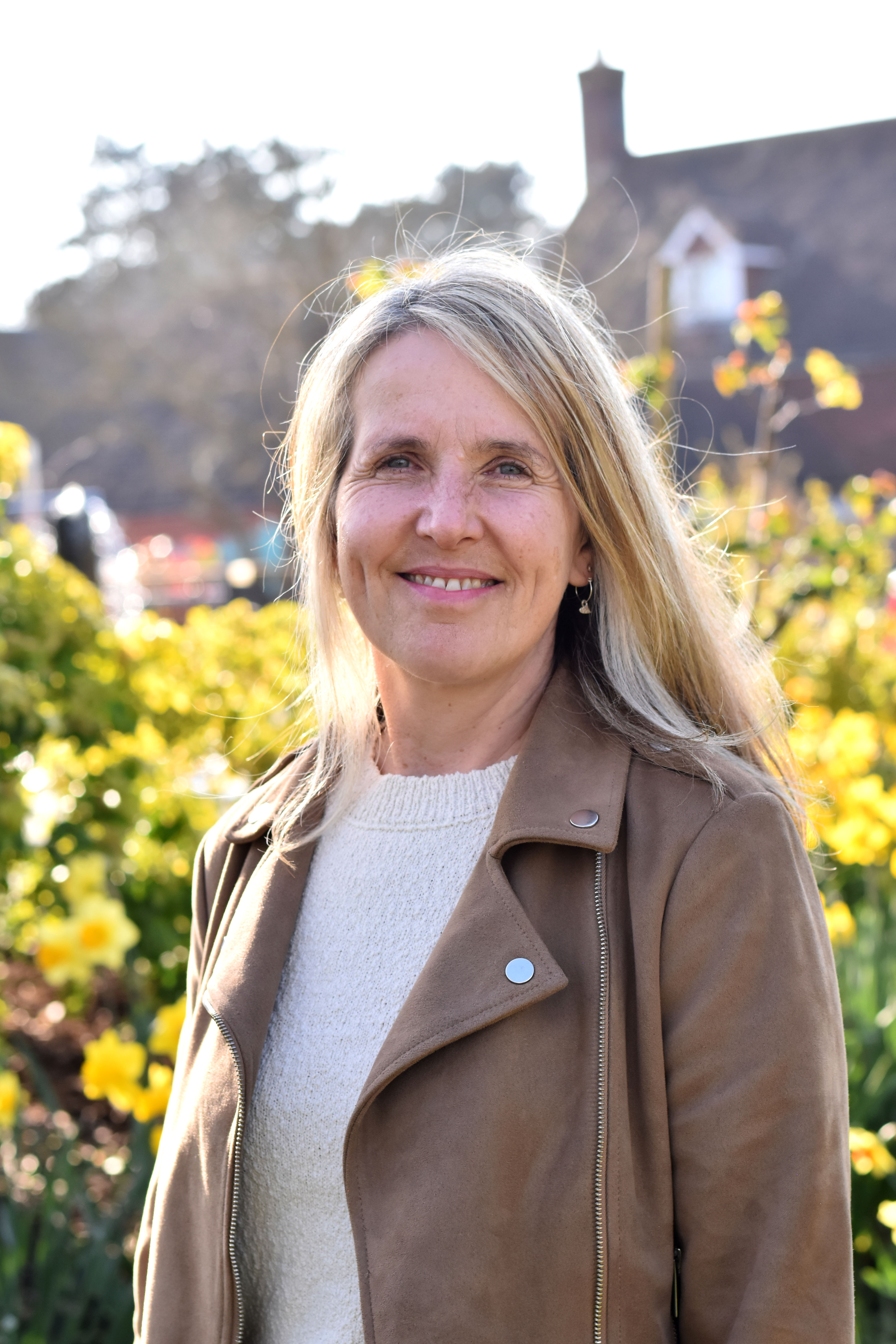 Trustee Heather Lucas smiles in sunny gardens, wearing a cream jumper and tan suede jacket.