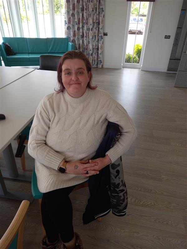 Person in white sweater and black trousers sitting on a chair in a room with wooden floor, large windows, and a green couch.