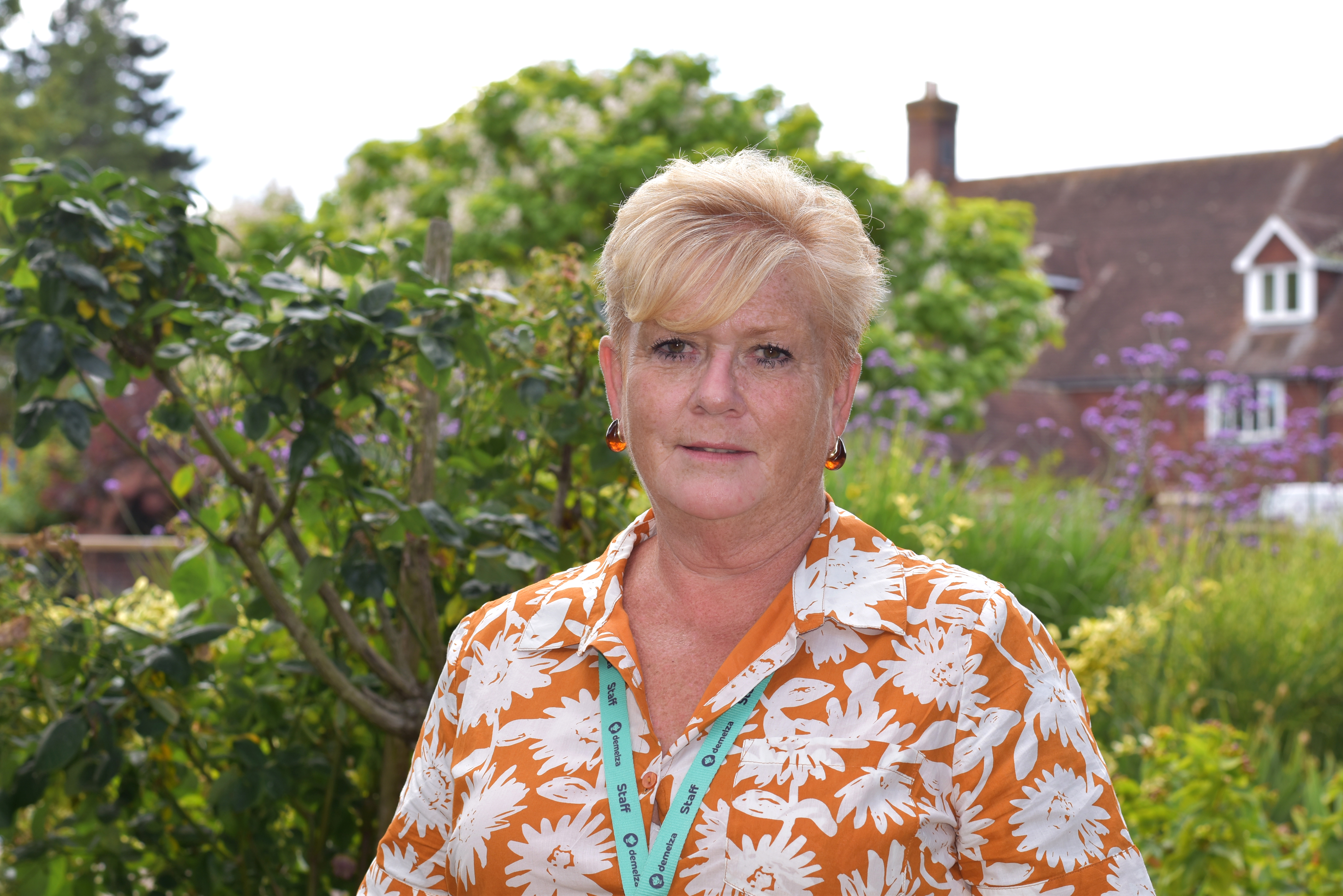 Demelza CEO Lavinia Jarrett stands in the hospice gardens, wearing a bright top and jewellery, smiling warmly.