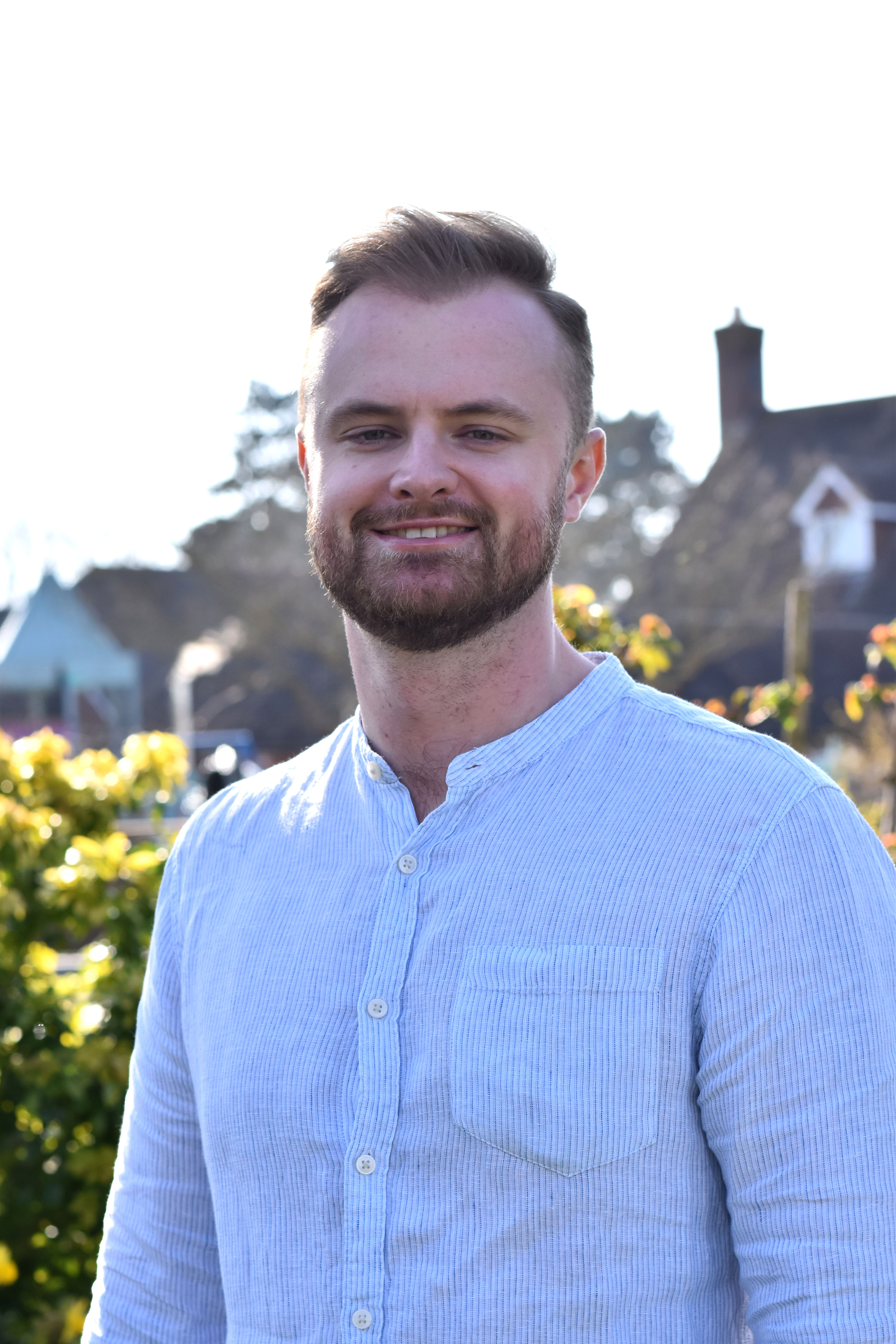 Trustee Rory Burns stands smiling in sunny gardens, wearing a pale blue shirt.