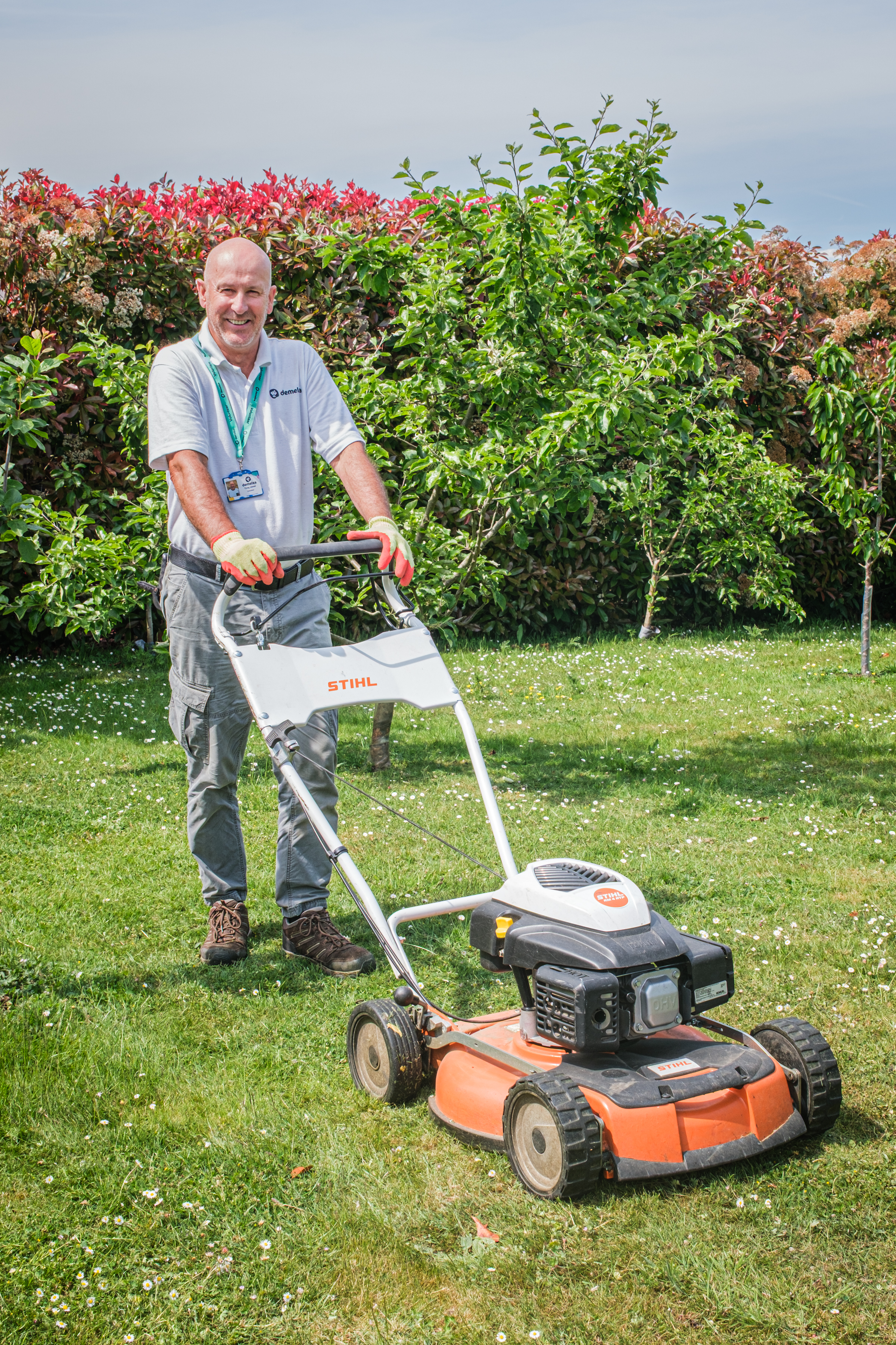 Man in white t-shirt with "demelza" wording pushing an orange and white STIHL lawnmower on a grassy lawn, with trees and bushes in the background.
