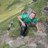 A blonde woman in a green shirt climbing up a grassy hill with mountains in the background