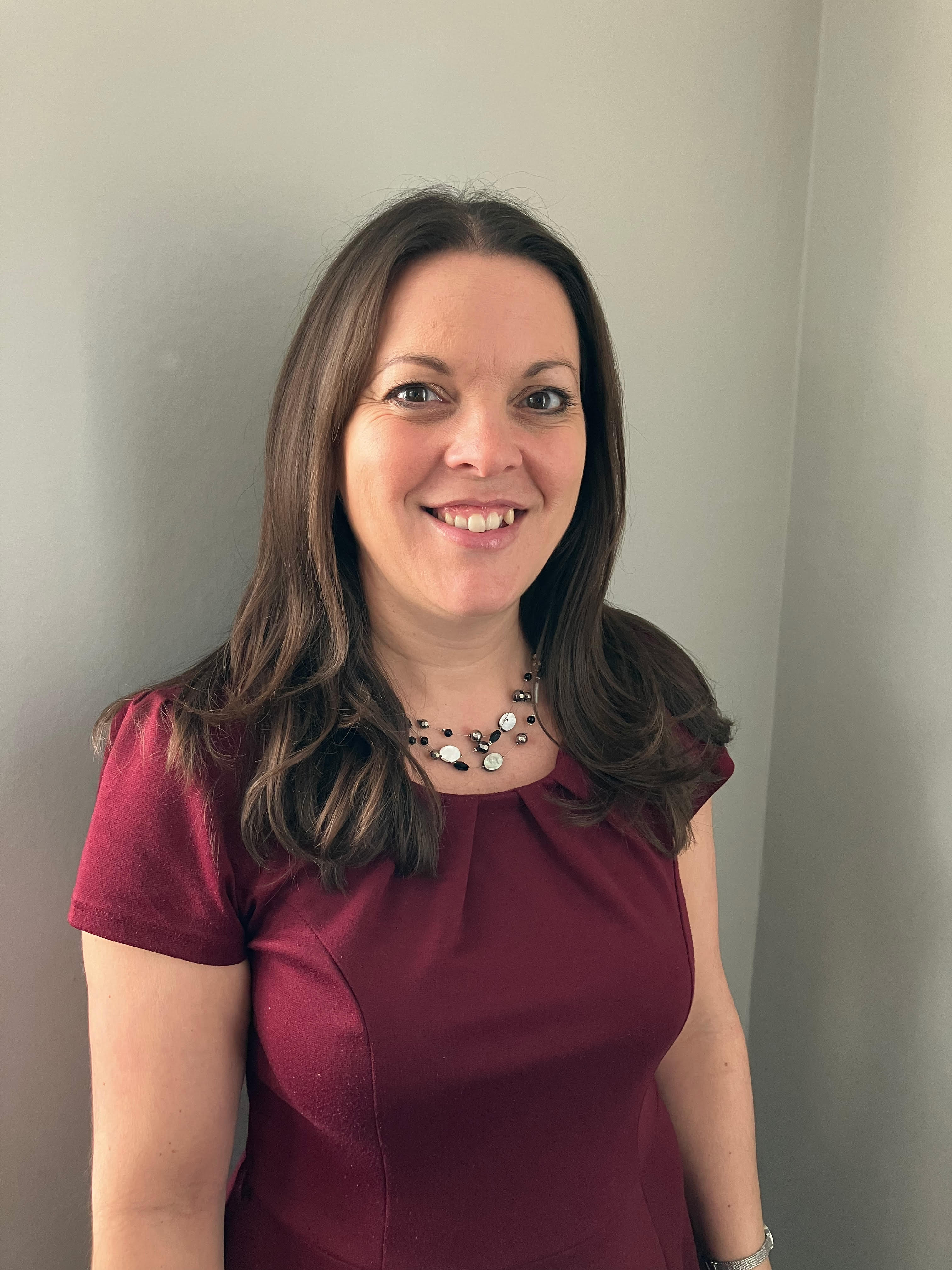 Trustee Natasha Smith smiles indoors, wearing a maroon smart dress and a pretty silver necklace.