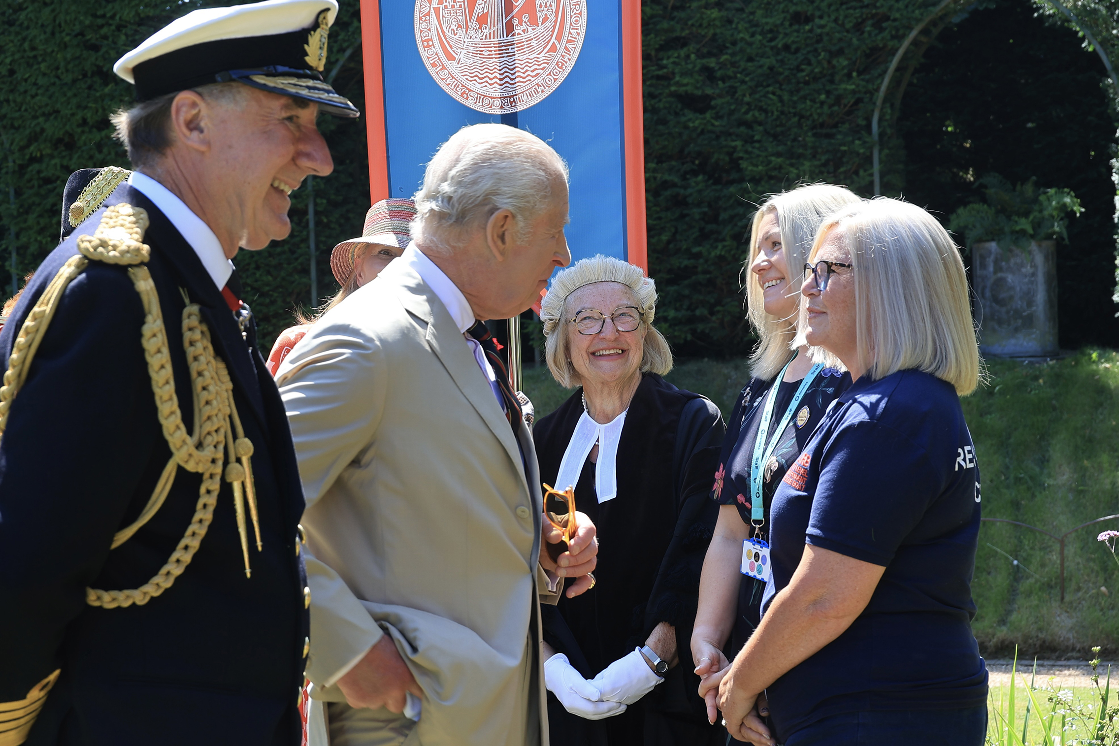 Group gathered outdoors near a blue and red banner with emblem. Includes man in military uniform and King Charles III in a beige suit.