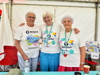 Three volunteers in bright clothes stand behind a table in a white marquee at the Mote Park Bubble Rush 2025 event.
