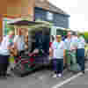 Five volunteer drivers in white uniforms stand by a minibus with its tail lift extended for wheelchair access.