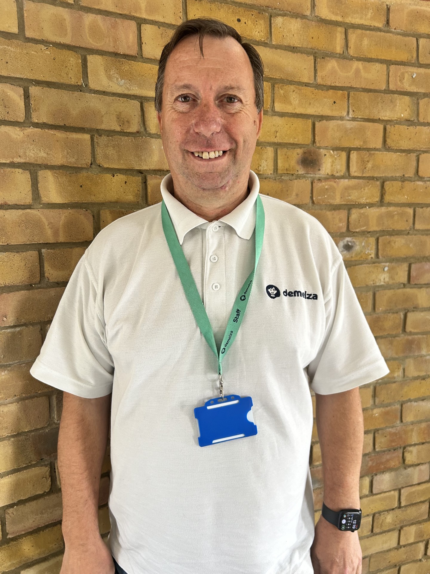 Person wearing a white polo shirt with "demelza" logo, standing in front of a light brown brick wall.