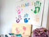 Colourful handprints and footprints labelled 'FAMILY' on a wall, with prints on wooden boards and cards. A vase of purple flowers sits nearby.