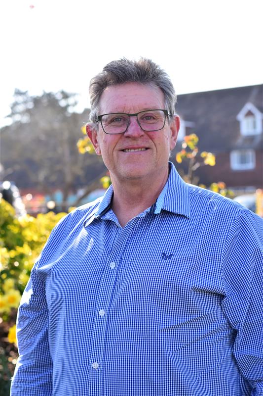 Trustee Dr Karl Holliman smiles in sunny gardens, wearing a blue checked long-sleeved shirt and black framed glasses.