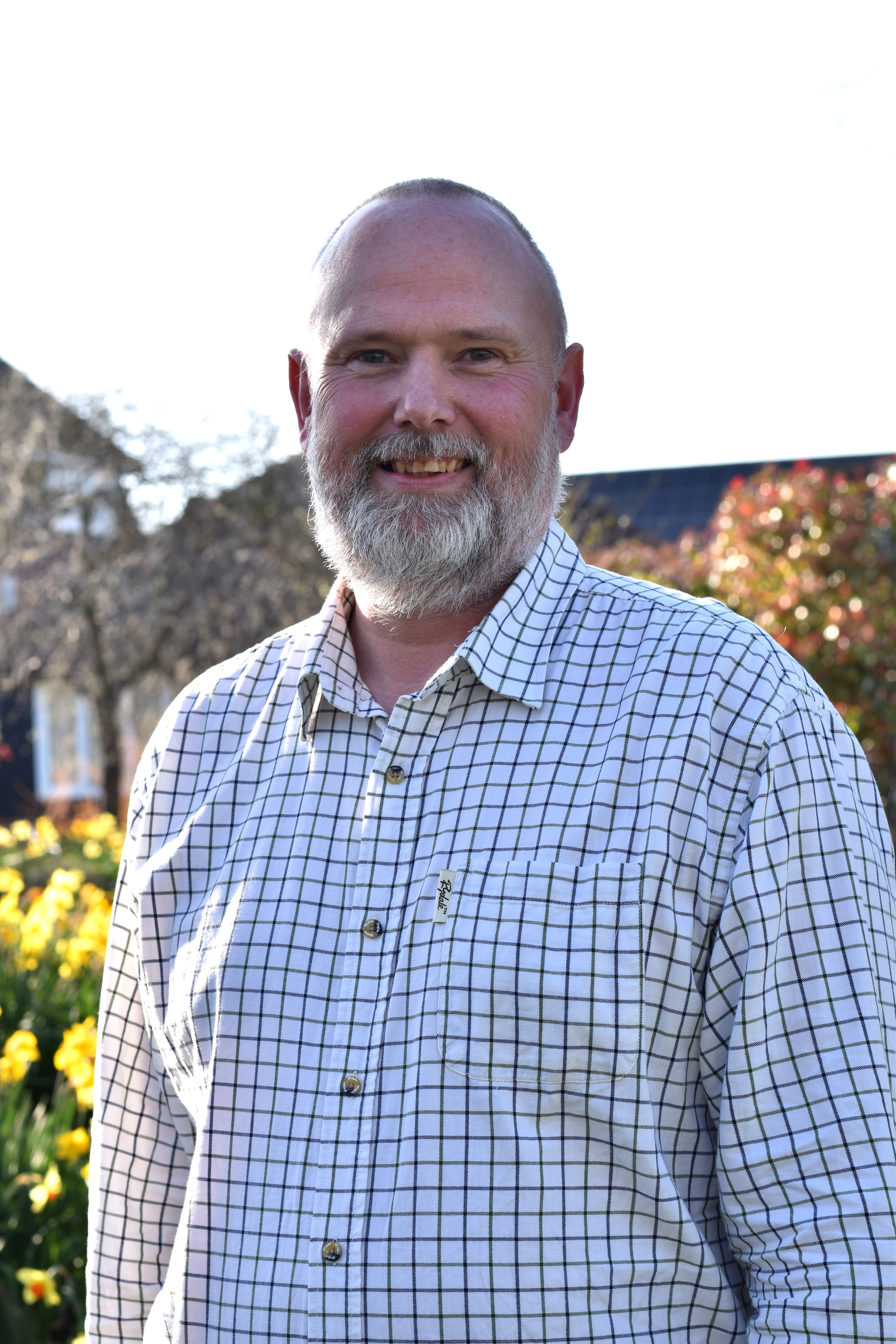 Chair of ACG Alex Parry-Jones stands smiling in sunny gardens, wearing a white and green checked shirt.