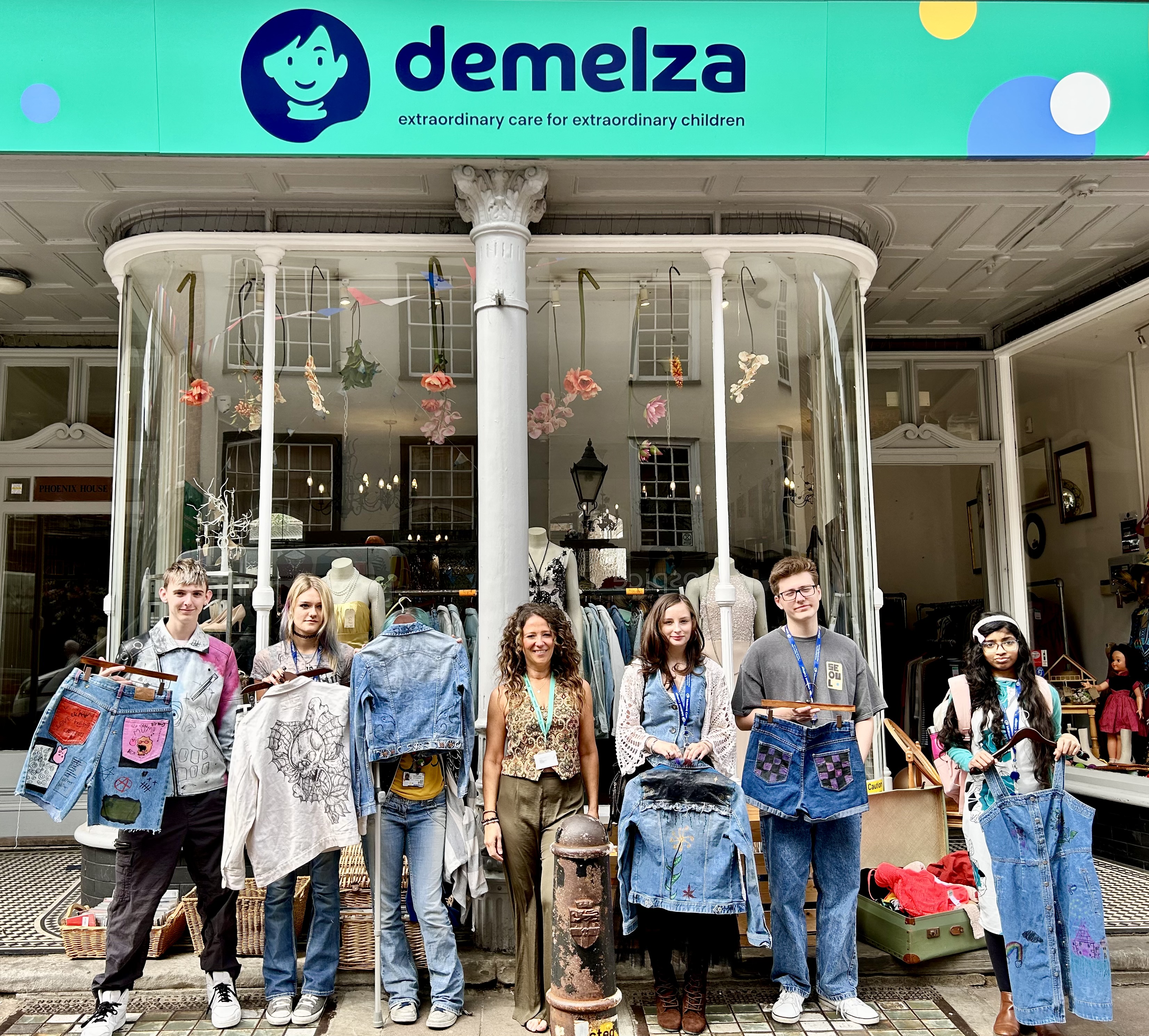 Group of people in front of Demelza charity shop holding denim clothing; shop sign reads "demelza extraordinary care for extraordinary children."