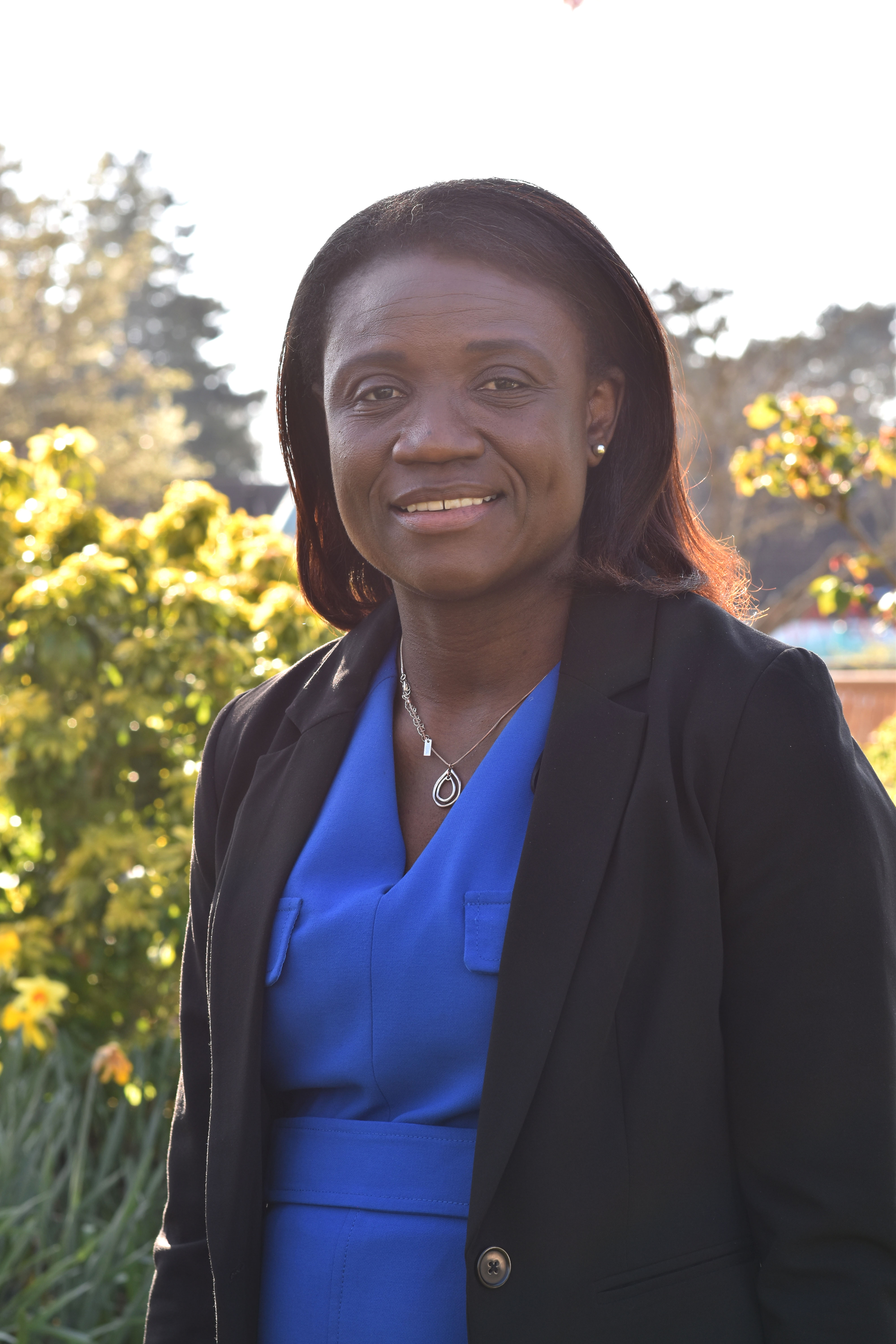 Trustee Dr Jenneh Tarawally stands in sunny gardens, wearing a smart blue dress and black blazer jacket