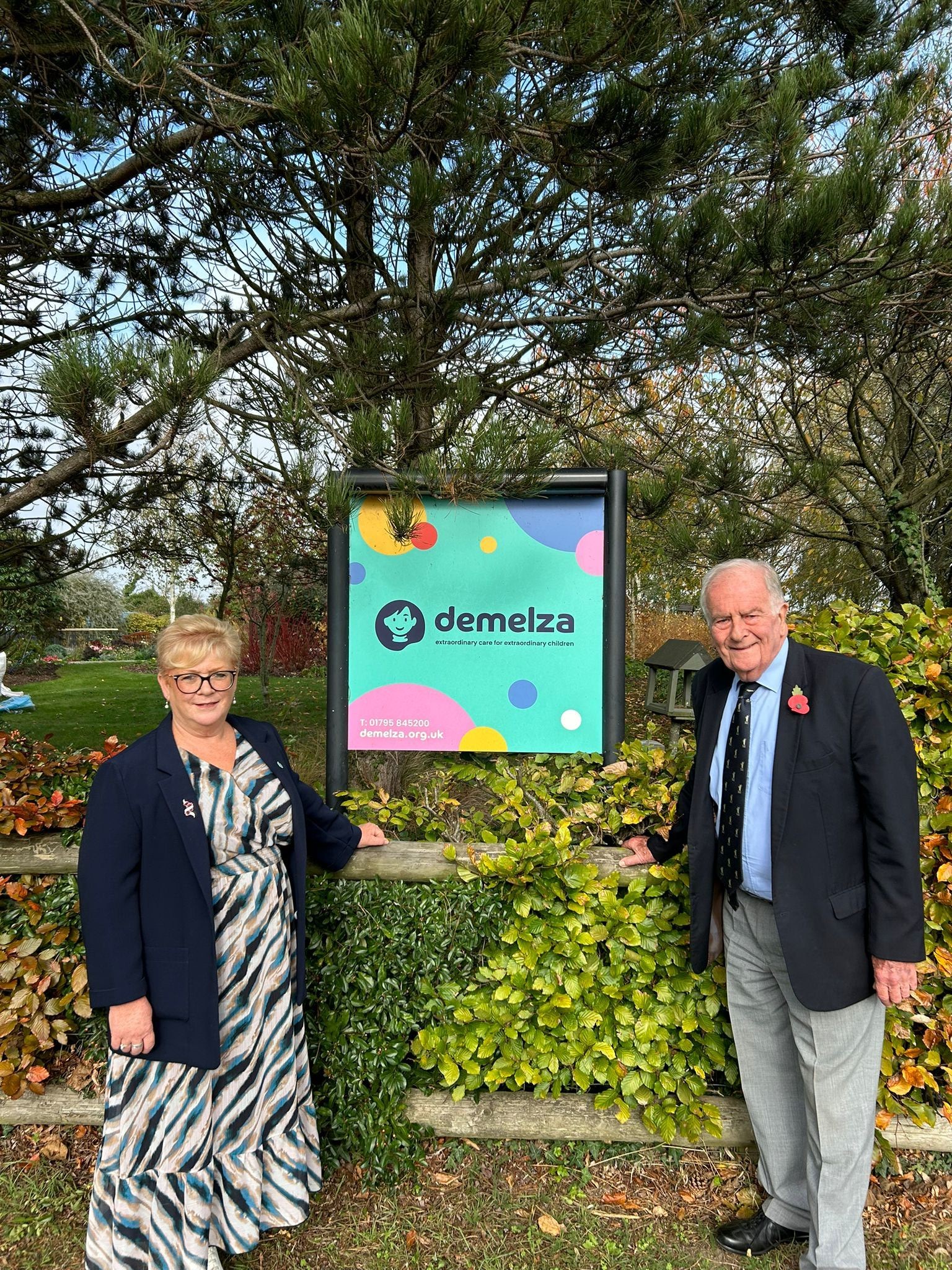 Two people standing outdoors beside a colourful "Demelza Hospice Care for Children" sign, surrounded by greenery and trees.