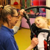 A nurse in a blue Demelza shirt, blonde hair and glasses, smiling towards a young girl with blonde hair in a wheelchair
