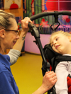 A nurse in a blue Demelza shirt, blonde hair and glasses, smiling towards a young girl with blonde hair in a wheelchair