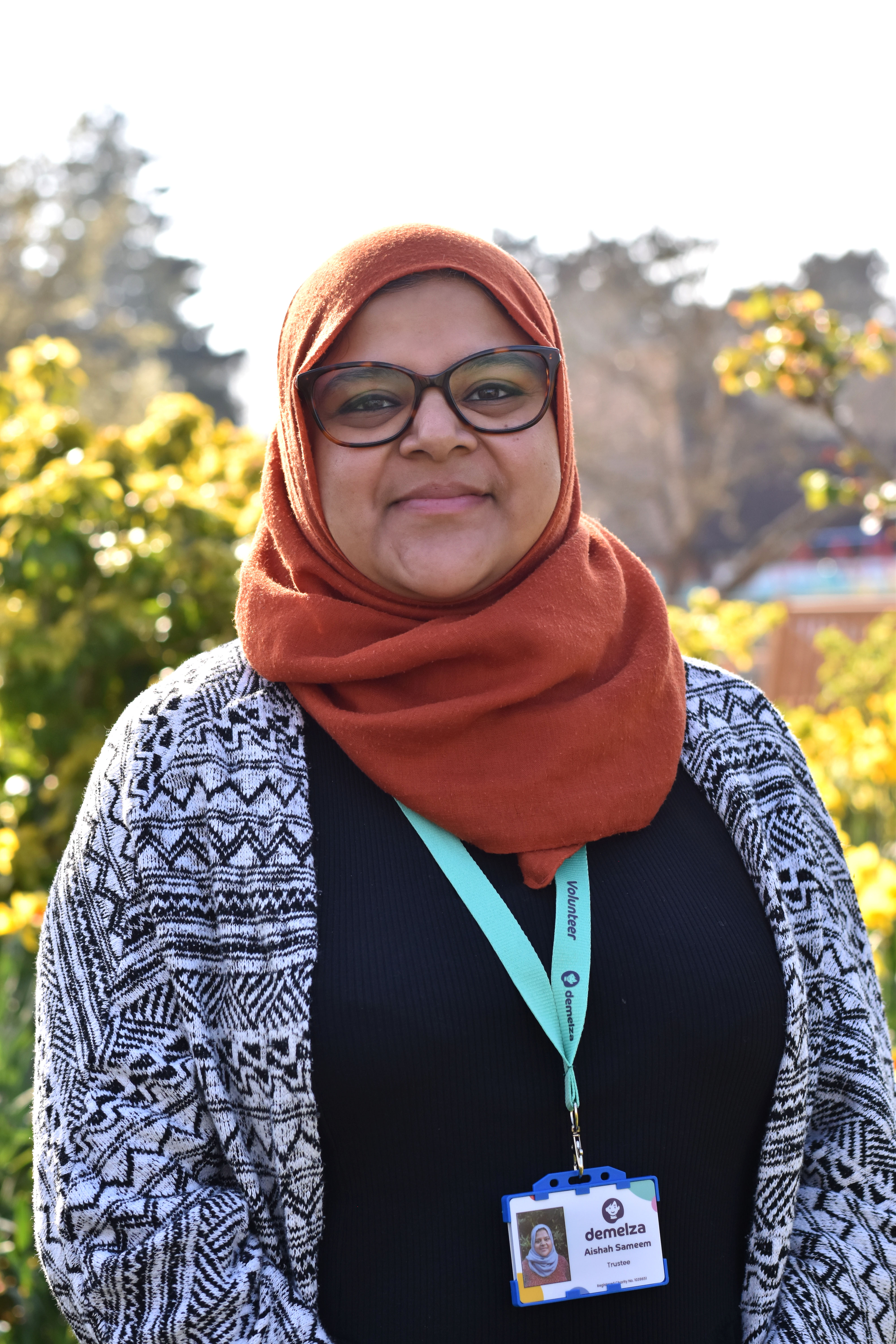 Trustee Aishah Sameem smiles in sunny gardens, wearing tortoise shell glasses, an orange headscarf and patterned cardigan.