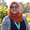 Trustee Aishah Sameem smiles in sunny gardens, wearing tortoise shell glasses, an orange headscarf and patterned cardigan.