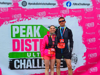 Two people in athletic gear with medals pose in front of a "PEAK DISTRICT ULTRA CHALLENGE" backdrop featuring hashtags and social media handles.