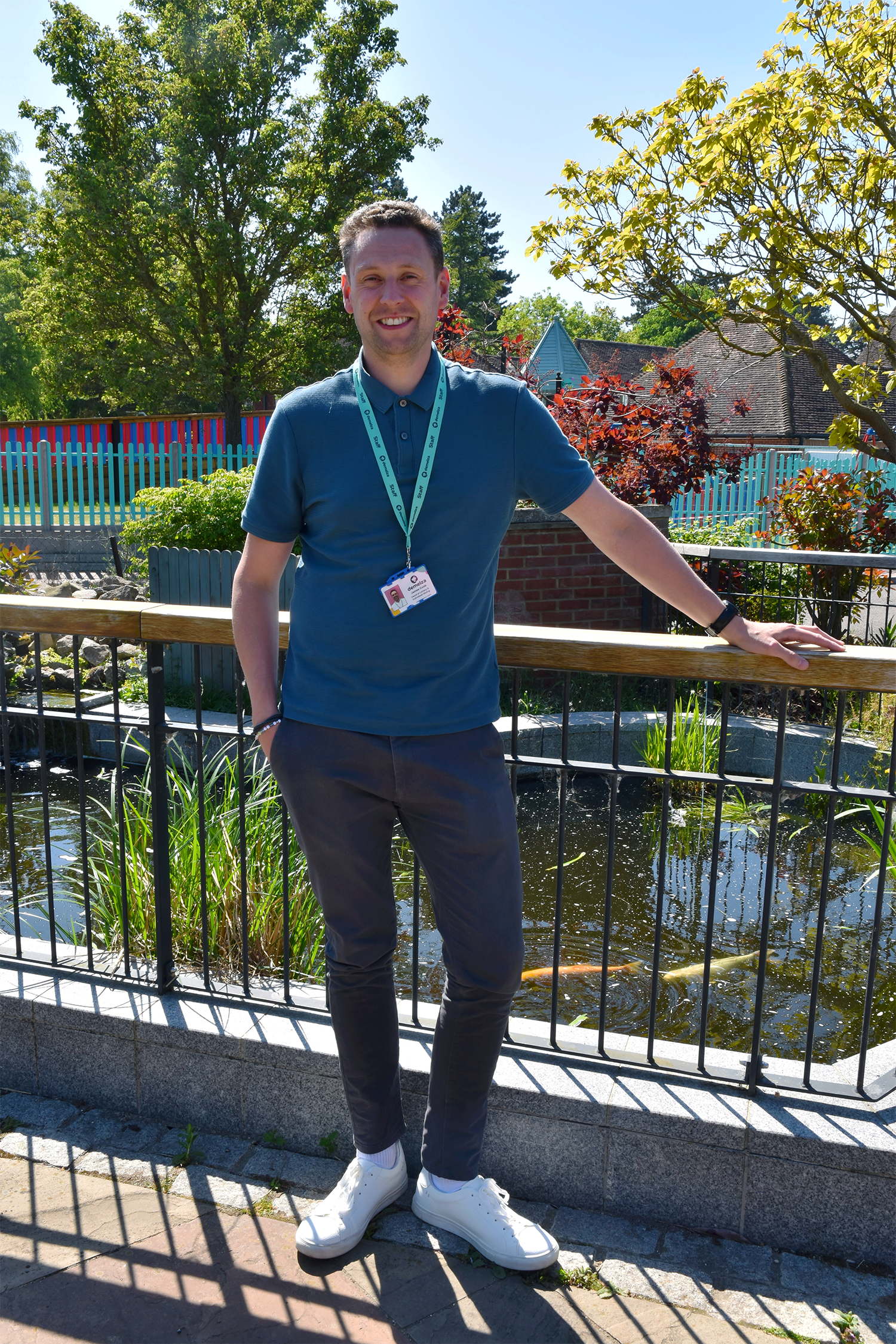 Man stands by a pond, wearing blue shirt, dark trousers, white shoes, and Demelza ID badge. Background includes trees, bushes, and a fence.