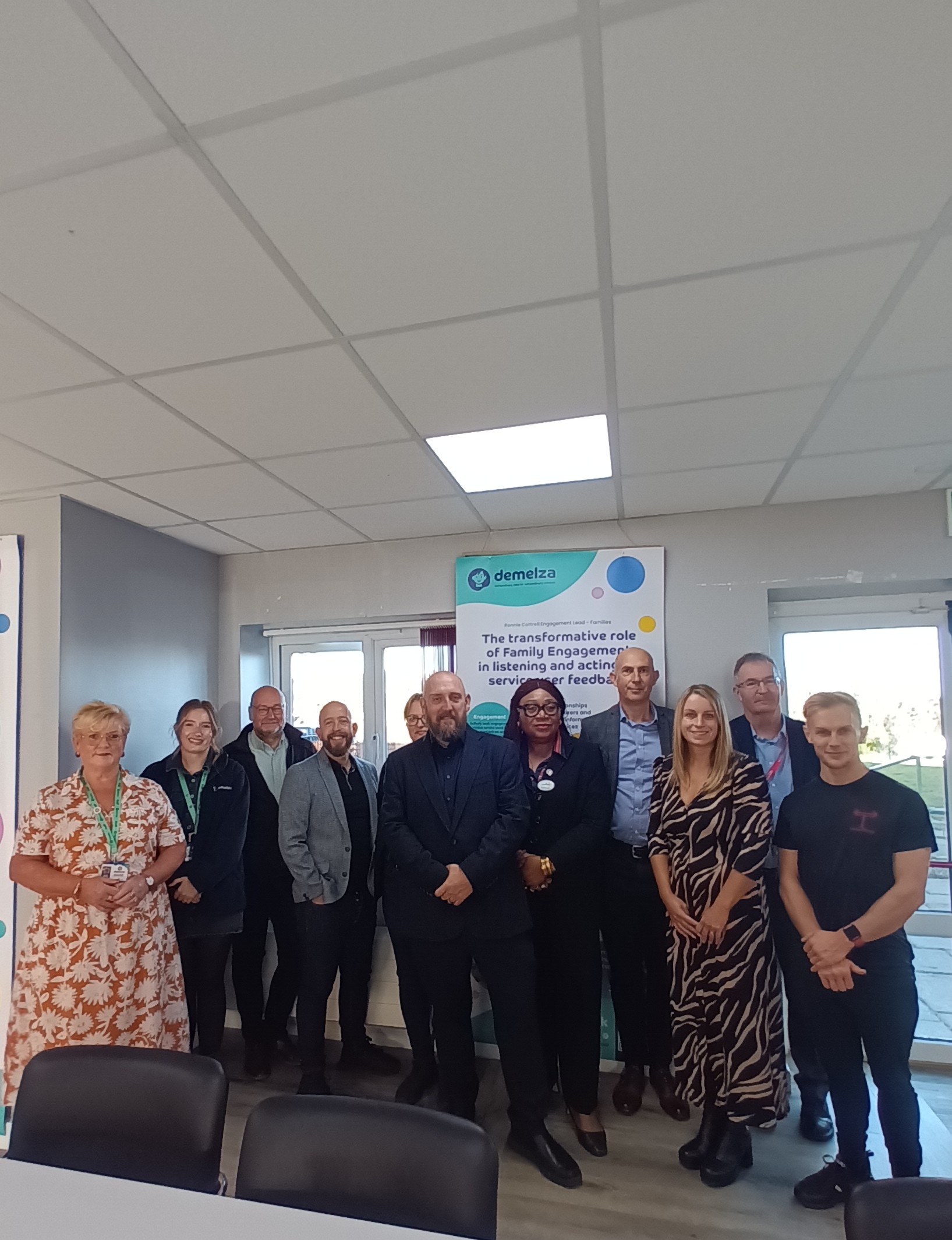 A group of people stand in a room with a white-tiled ceiling. Behind them is a banner that reads, "The transformative role of Family Engagement in listening and acting on service user feedback," with the "demelza" logo at the top.