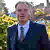 Chair of Safeguarding Assurance Committee Dr Richard Patey stands in sunny gardens wearing a smart navy blue three-piece suit and tie.