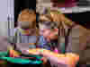 A woman with blonde hair and glasses smiling while helping a young boy on a tablet play the piano