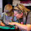A woman with blonde hair and glasses smiling while helping a young boy on a tablet play the piano