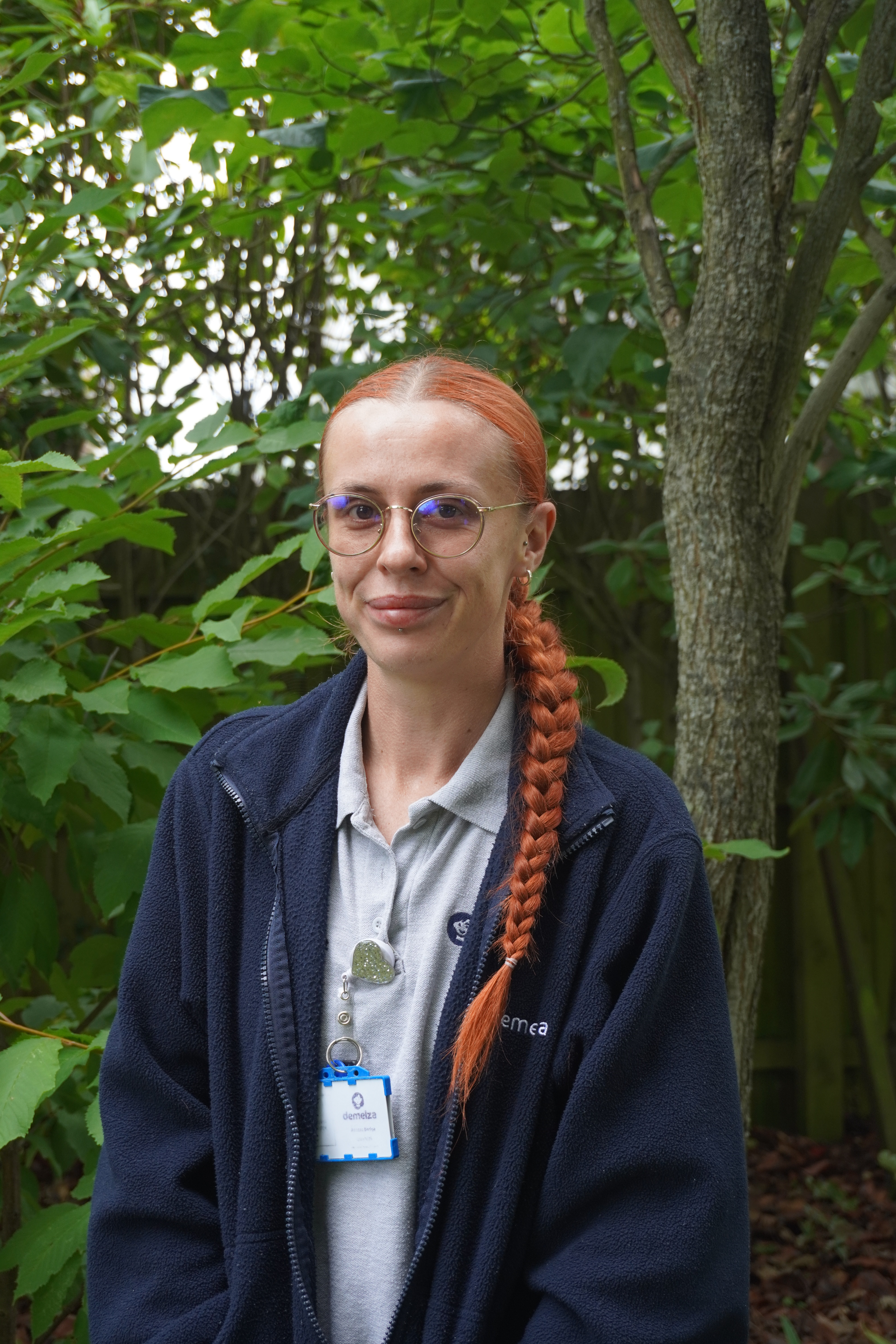 A person with long red braided hair stands outdoors in front of green foliage and trees. They are wearing a blue jacket over a light-colored shirt and have a Demelza identification badge hanging from their neck.