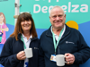 A man with white hair and a woman with brown hair holding mugs while smiling at the camera.