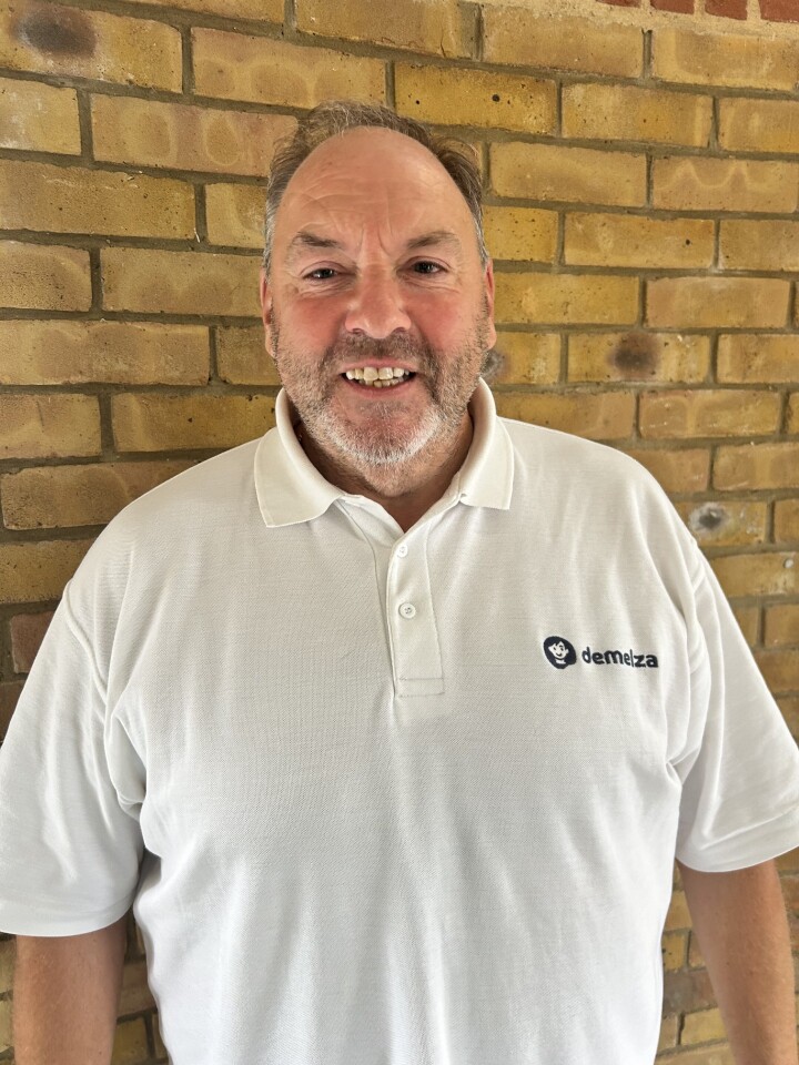Person wearing a white polo shirt with "demelza" logo, standing in front of a light brown brick wall.