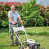 Smiling male volunteer pushes a lawn mower across a sunlit garden, trimming grass under clear blue skies.