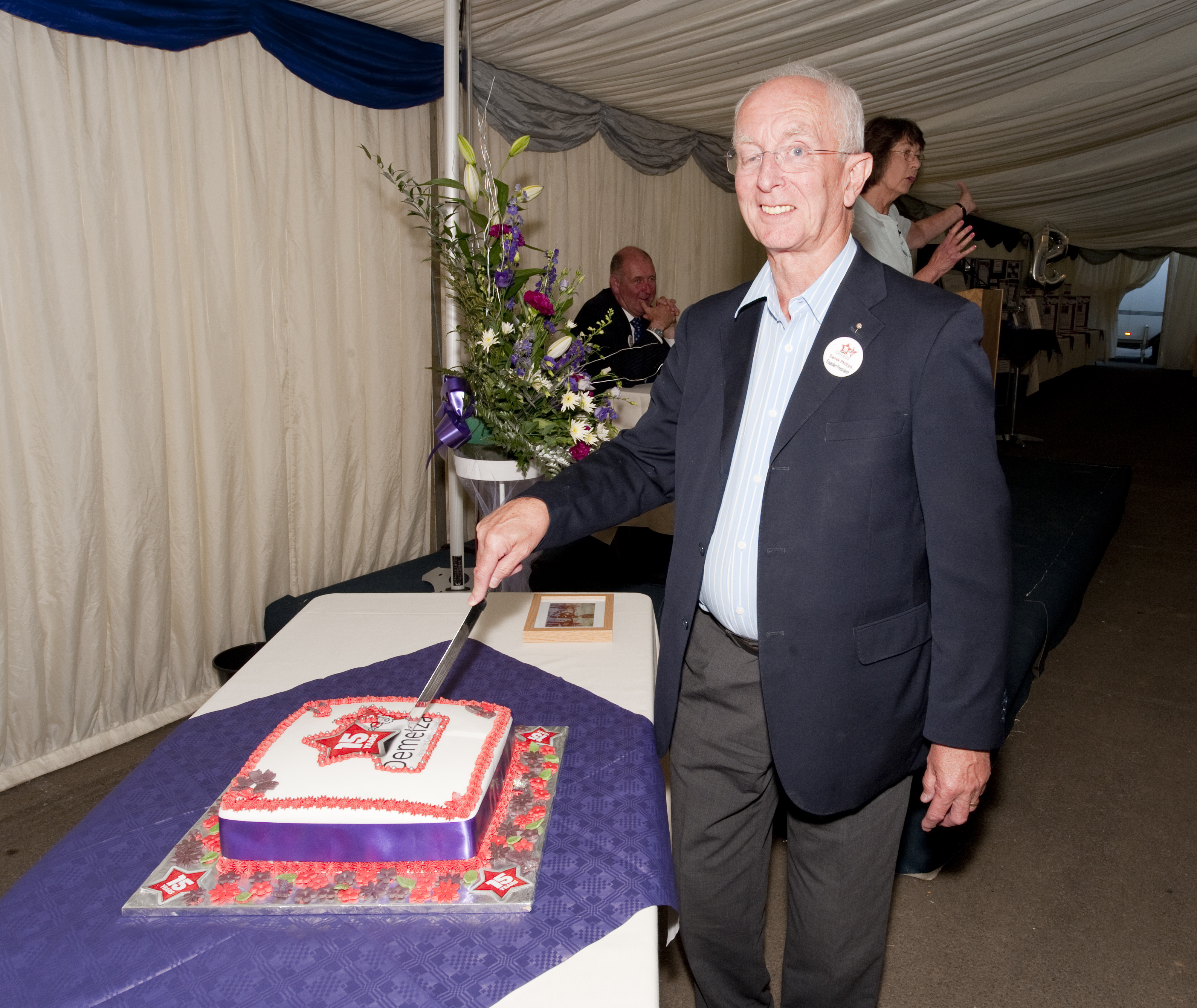 Man in a suit cutting a decorated cake on a table inside a tent, with flowers and other people visible in the background.