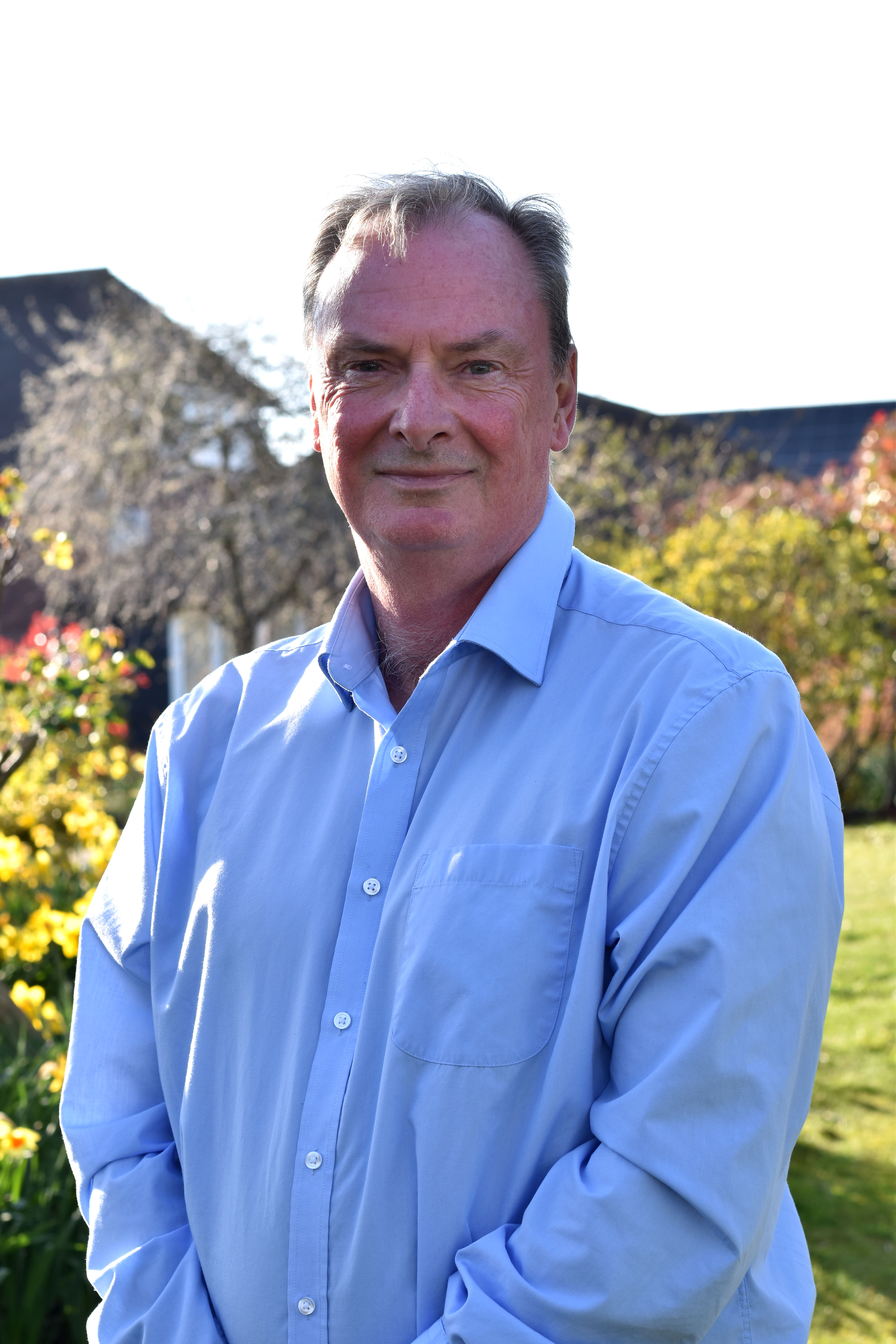 Chair Sean Reynolds CB CBE DFC stands smiling in sunny gardens, wearing a long-sleeved pale blue shirt.