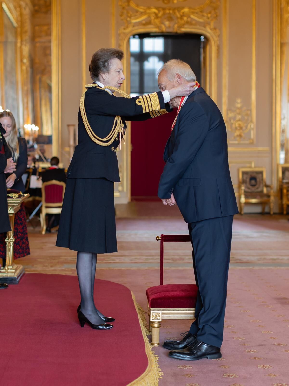 Person in navy uniform places medal on man in suit during formal ceremony in ornate room with gold decor and red and gold  chairs.