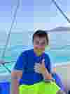 A child in a blue shirt and yellow shorts on a boat smiling towards the camera with a thumbs up