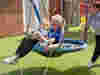 Demelza nurse and child sitting on a circular swing, with another adult standing nearby on a playground with artificial grass.